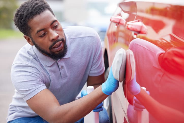 Man polishing a red car with a cloth, wearing gloves and a polo shirt, focused on detailing the car's surface.